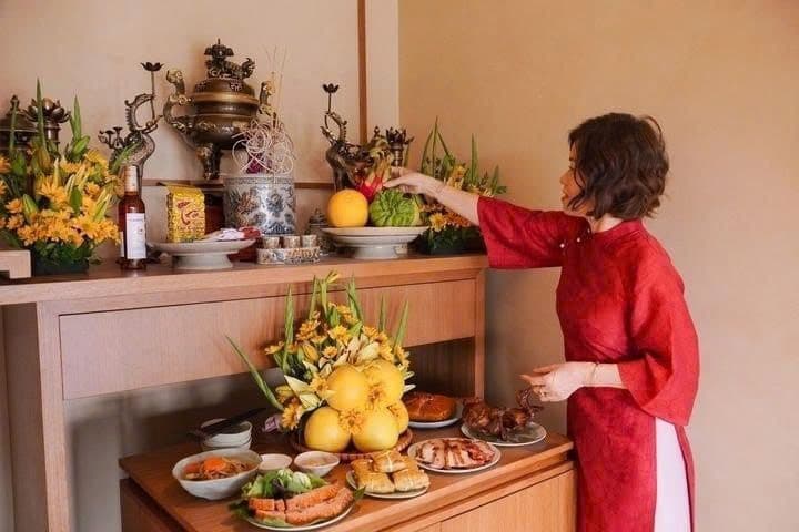 Người phụ nữ mặc áo dài đỏ đang chuẩn bị mâm cỗ cúng Ông Công Ông Táo trên bàn thờ.