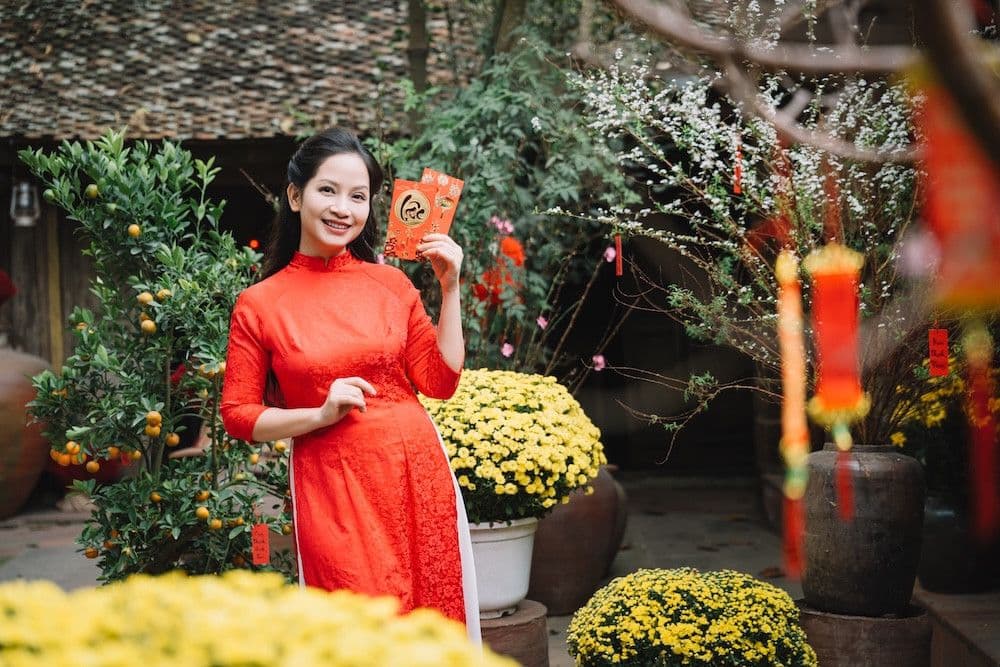 Người phụ nữ mặc áo dài đỏ cầm bao lì xì, mỉm cười trong không khí Tết vui tươi, tượng trưng cho niềm vui nhạc xuân.