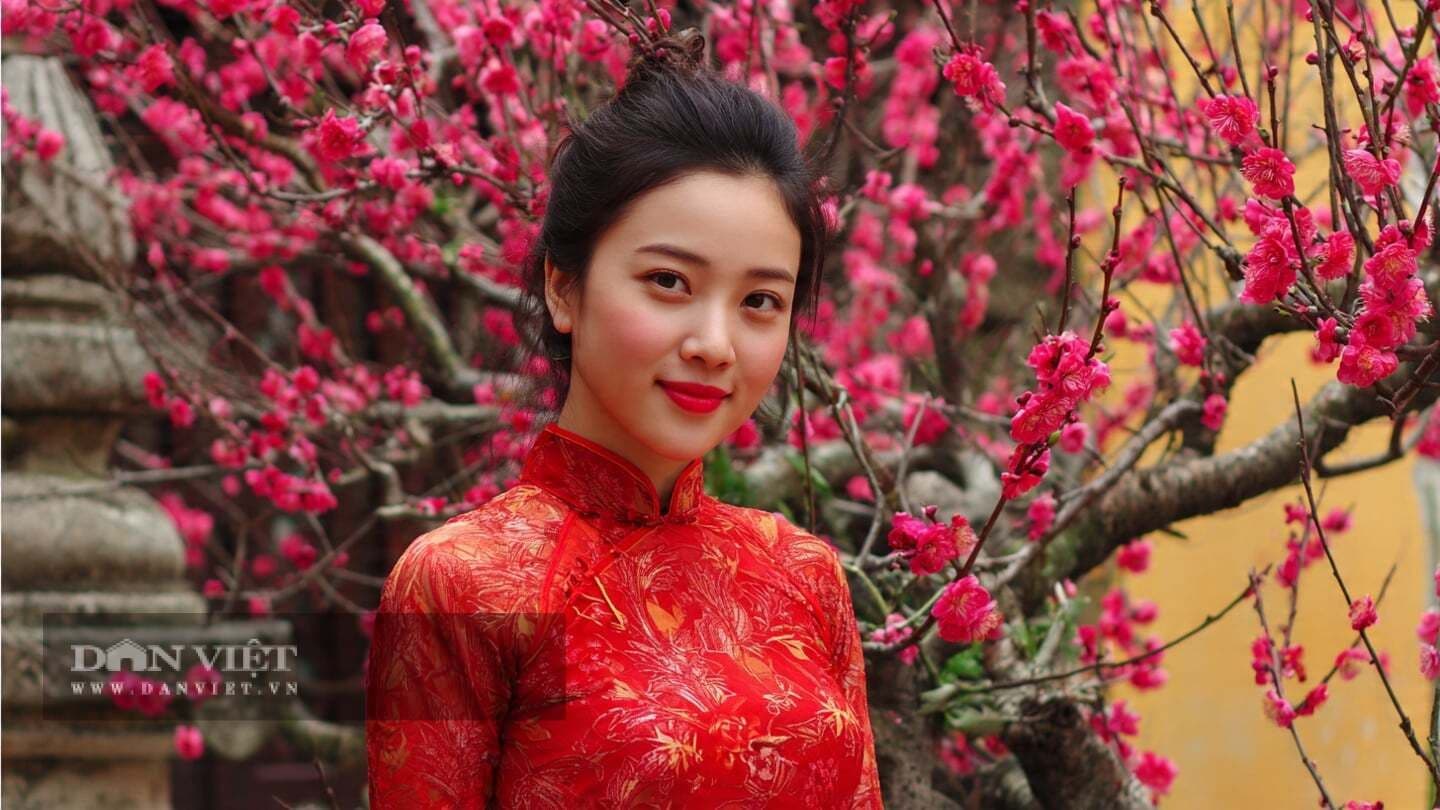 Người phụ nữ Việt Nam trong áo dài đỏ tươi cười giữa vườn hoa đào, tượng trưng cho may mắn và khởi đầu tốt lành.
