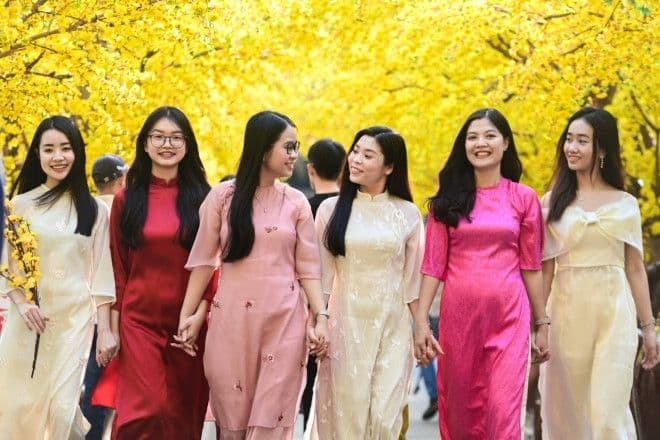 Nhóm phụ nữ Việt Nam trong trang phục áo dài truyền thống dạo bước dưới hàng cây hoa mai vàng rực rỡ, thể hiện nét đẹp văn hóa ngày Tết.