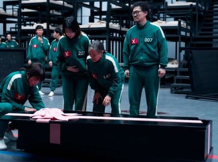 Những người chơi Squid Game trong bộ đồ xanh lá cây đứng quanh một chiếc quan tài, thể hiện sự tàn khốc của trò chơi.