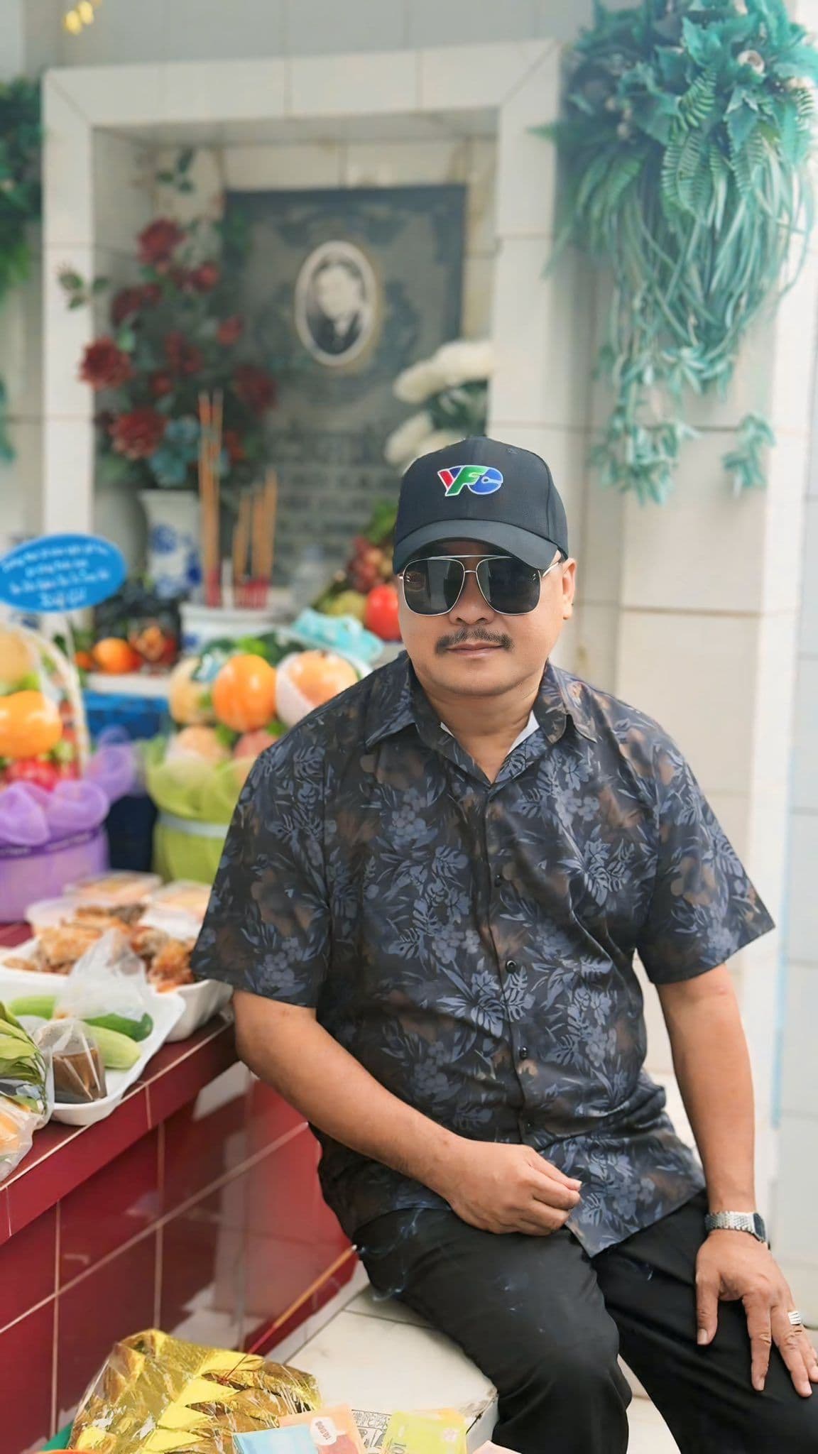 Phước Sang đội mũ và kính râm, ngồi tại mộ Lê Công Tuấn Anh, thể hiện sự tưởng nhớ bền bỉ.