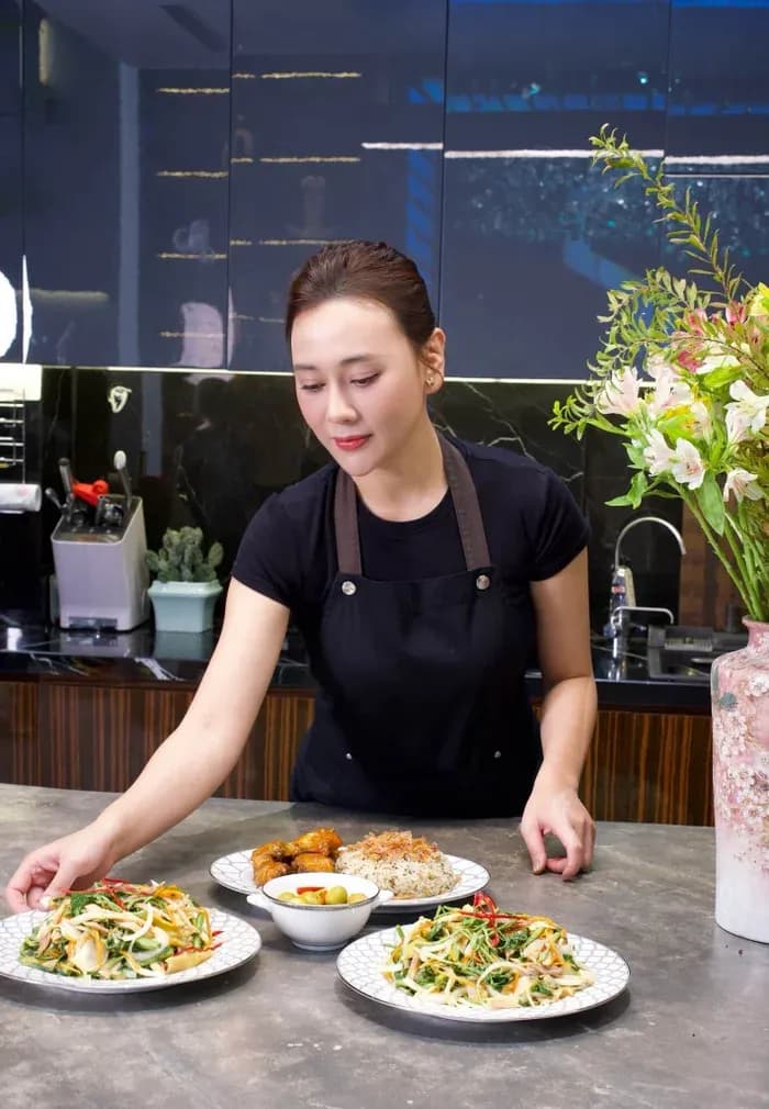 Phương Oanh trong bếp với áo phông đen và tạp dề, đang bày biện các món ăn tự chế biến như xôi gà rô-ti và nộm gà xé phay.
