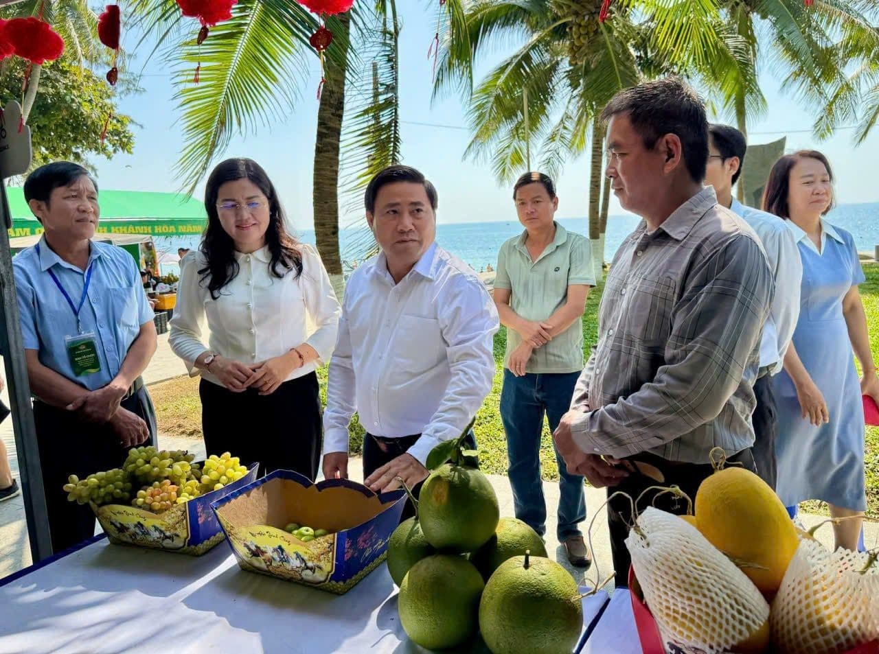 Quan chức và nông dân Khánh Hòa kiểm tra các sản phẩm nông sản tươi như bưởi, xoài và nho tại một gian hàng bên bờ biển, minh họa hành trình từ vườn ra chợ.