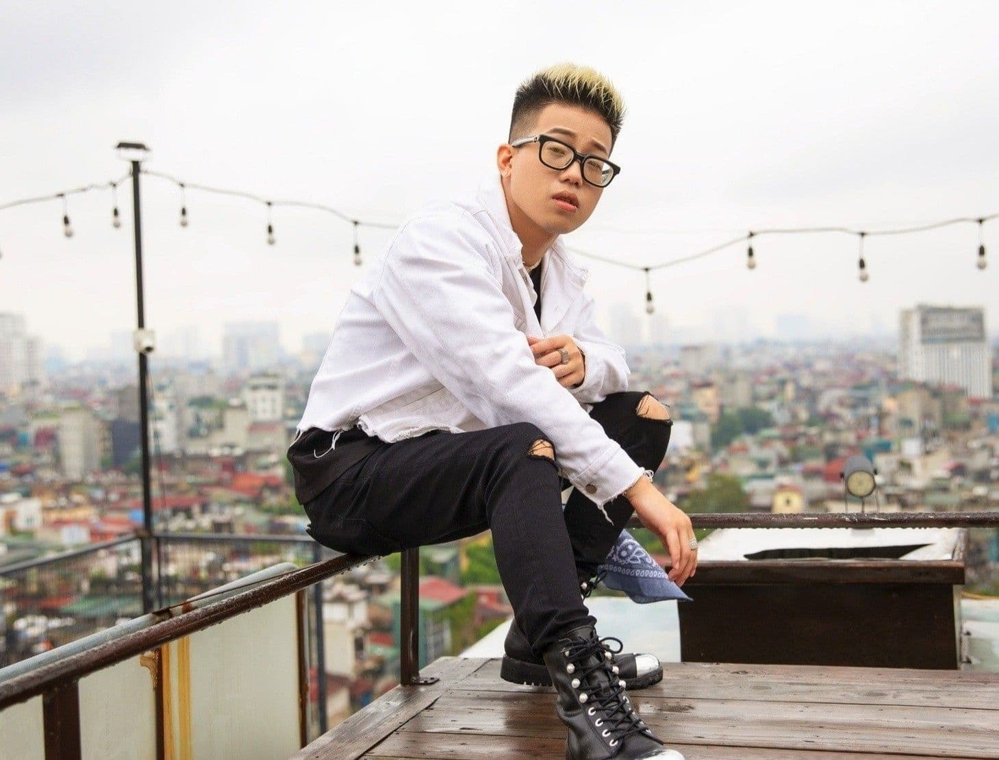 RichChoi, rapper người Việt, ngồi trên sân thượng với áo khoác trắng và kính, nhìn thẳng vào ống kính.
