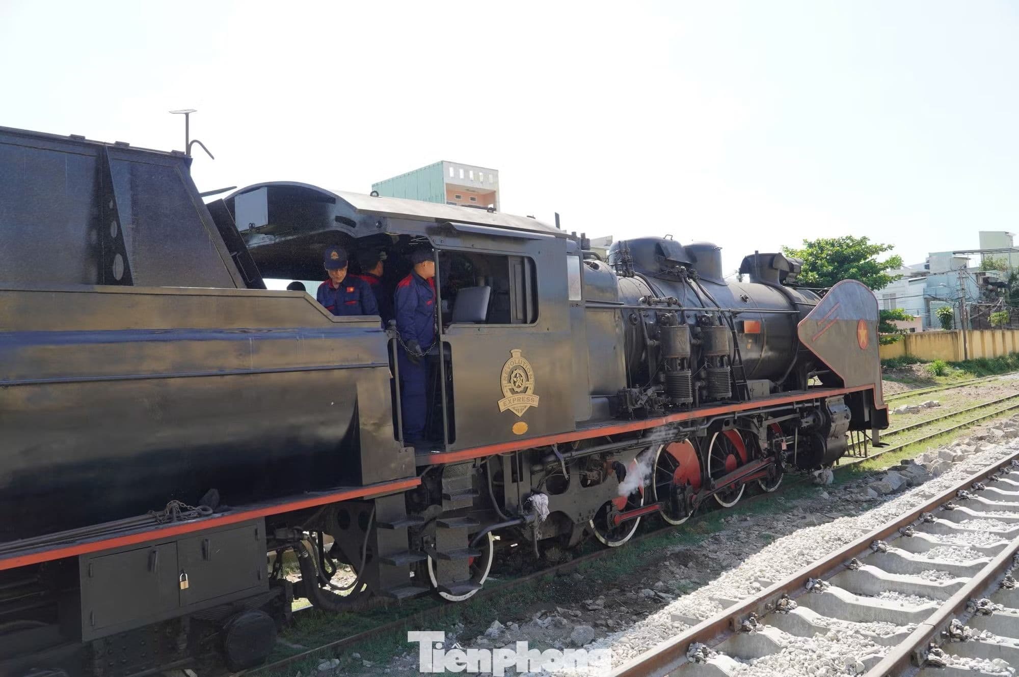 Đầu máy hơi nước cổ điển đang di chuyển trên đường ray, với hai người trong buồng lái, gợi nhớ ký ức về ngành đường sắt Việt Nam giữa đô thị hiện đại.